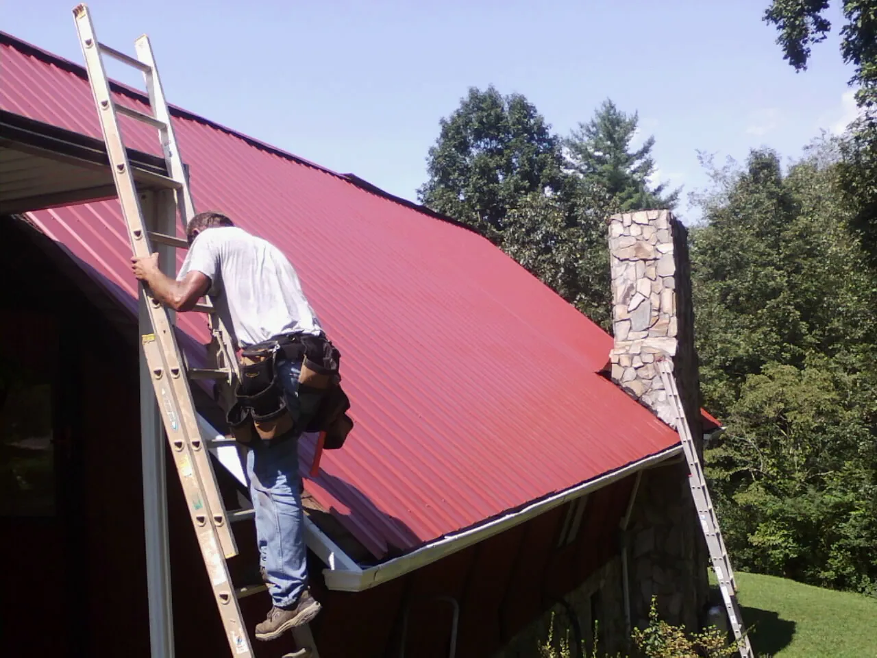 Roofer on a ladder beside a red metal roof and stone chimney — Soffit & Fascia Repair by Roofing Brothers in Haskell